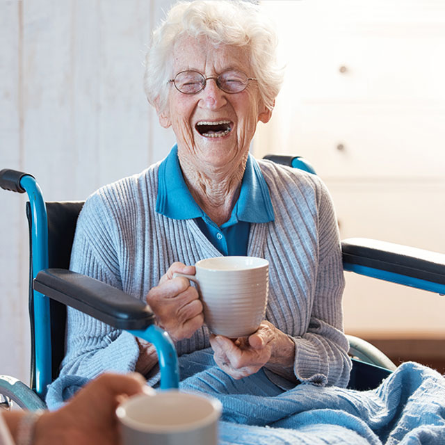 Eine ältere Dame in einem Rollstuhl lacht herzlich und hält eine Tasse, die entspannte und freudige Atmosphäre während der Betreuung durch den Pflegedienst AML ausdrückt.