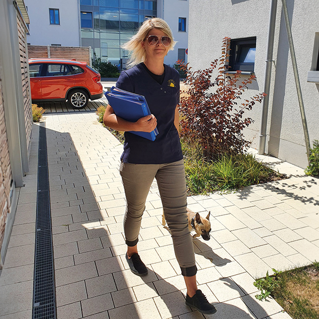 Eine Pflegerin von Pflegedienst AML geht mit einer Mappe und einem Hund an der Leine einen Weg entlang, was die Kostenberatung und individuelle Betreuung symbolisiert.